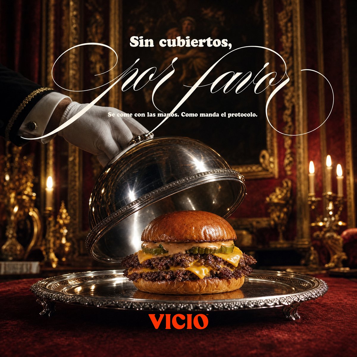 Anuncio de Vicio: una hamburguesa servida bajo campana de plata en un salón barroco. «Sin cubiertos, por favor».